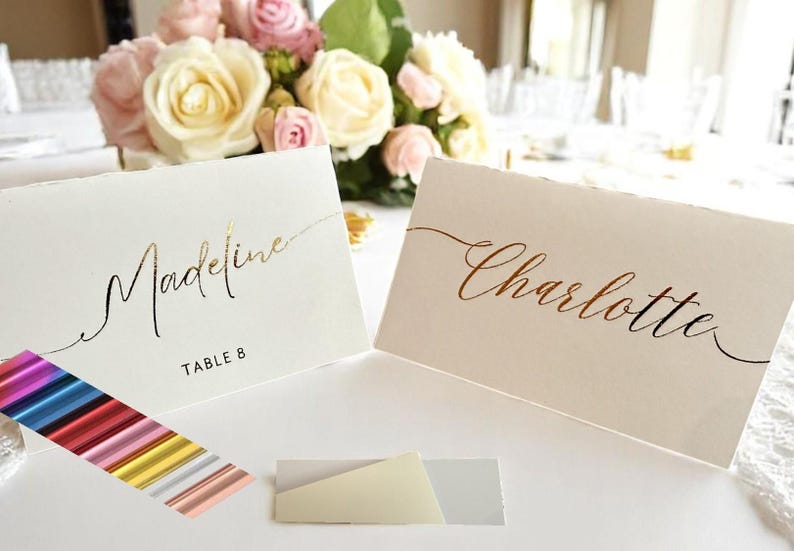 May include: Two white place cards with gold and copper foil lettering. The cards read "Madeline, Table 8" and "Charlotte".
