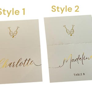 May include: Two styles of place cards for a wedding or special event. Style 1 features a gold foil design of two champagne glasses with the name "Charlotte" written in gold foil. Style 2 features the same champagne glass design with the name "Madeline" and "Table 8" written in gold foil.