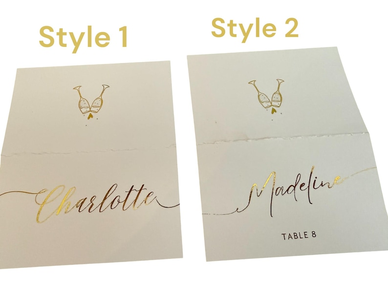 May include: Two styles of place cards for a wedding or special event. Style 1 features a gold foil design of two champagne glasses with the name "Charlotte" written in gold foil. Style 2 features the same champagne glass design with the name "Madeline" and "Table 8" written in gold foil.