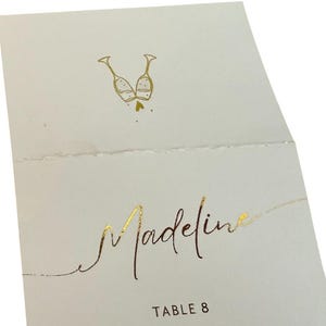 May include: A white place card with gold foil lettering. The card has a design of two champagne glasses clinking together. The text on the card reads "Madeline" and "Table 8".