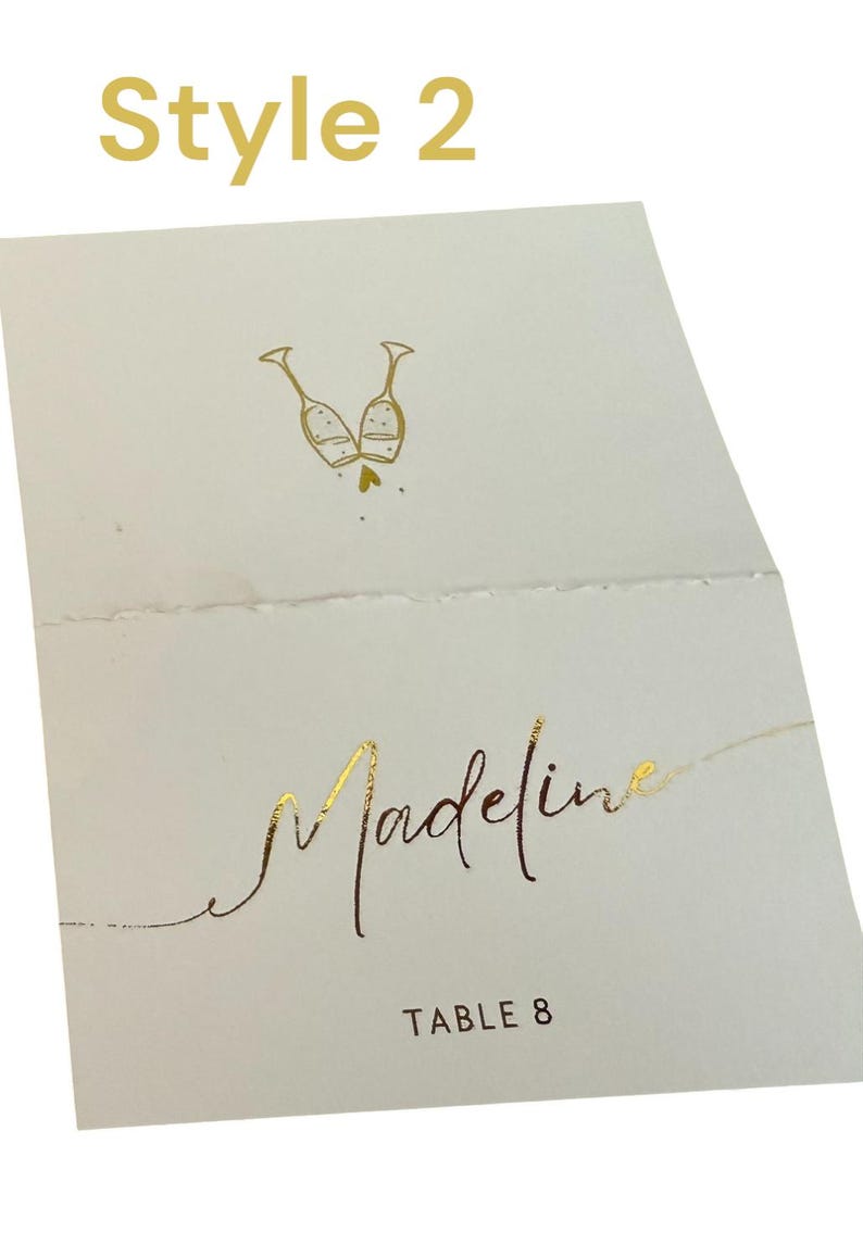 May include: A white place card with gold foil lettering. The card has a design of two champagne glasses clinking together. The text on the card reads "Madeline" and "Table 8".