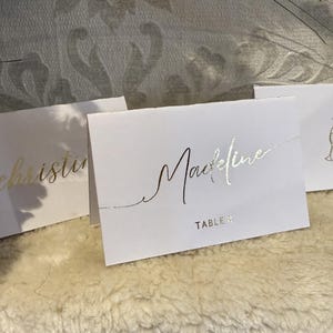 May include: Three white place cards with gold lettering. The first card says "christine", the second card says "Maxine" and "TABLE 4", and the third card has a gold illustration of two champagne glasses with a heart.