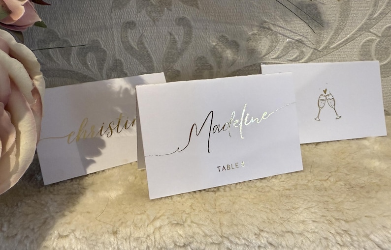 May include: Three white place cards with gold lettering. The first card says "christine", the second card says "Maxine" and "TABLE 4", and the third card has a gold illustration of two champagne glasses with a heart.
