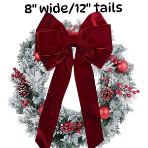 May include: A snowy evergreen wreath with red berries and baubles, decorated with a large, red velvet bow with 8-inch wide and 12-inch long tails.