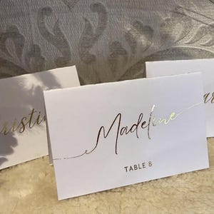 May include: Three white place cards with gold lettering. The cards read "Christine", "Madeline Table 8", and "Charlotte".
