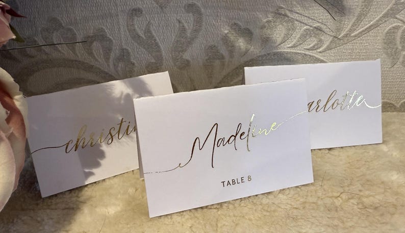 May include: Three white place cards with gold lettering. The cards read "Christine", "Madeline Table 8", and "Charlotte".