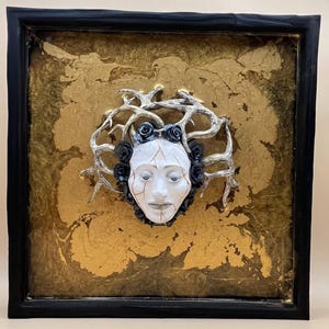 May include: Framed artwork with a white face, cracked and decorated with black roses and a silver crown. The face is set against a gold and brown textured background, within a black frame. The artwork has a surreal, artistic theme.