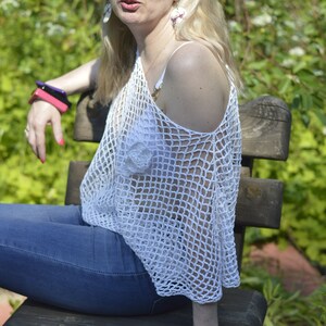 May include: A white crochet top with a loose, open-weave design. The top has a single strap and a wide, flowing neckline. The model is wearing blue jeans.