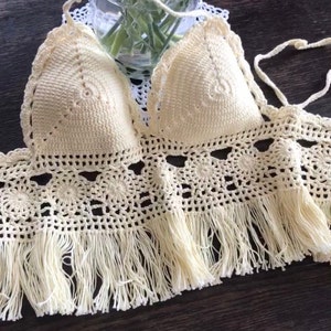 May include: A crocheted crop top with a delicate, lacy design and a fringe trim. The top is a light cream color and has a halter style.