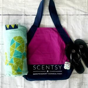 May include: A pink tote bag with navy blue straps and trim. The bag has the text "Scentsy" and "Independent Consultant" printed on the front. A green and blue geometric cactus print beach towel and black flip flops are next to the bag.