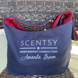 May include: A navy blue canvas tote bag with a white embroidered logo that reads "Scentsy" with a star and the text "Independent Consultant Amanda Brown".