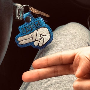 May include: A blue and white key chain with the word "UNAGI" embroidered on it. The keychain is shaped like a hand making the peace sign.