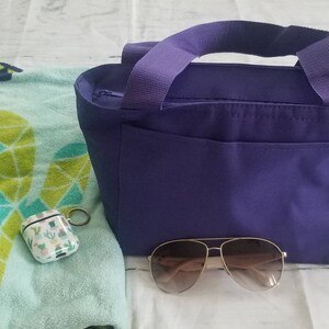 Puede incluir: Una bolsa de mano de color morado con cierre de cremallera y un bolsillo frontal. La bolsa est&aacute; sobre una superficie blanca junto a una toalla de playa verde y azul, una funda blanca con dise&ntilde;o de cactus y unas gafas de sol marrones.