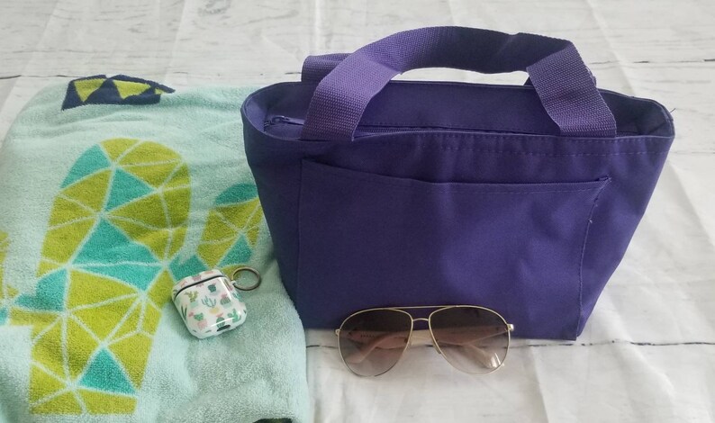 Puede incluir: Una bolsa de mano de color morado con cierre de cremallera y un bolsillo frontal. La bolsa est&aacute; sobre una superficie blanca junto a una toalla de playa verde y azul, una funda blanca con dise&ntilde;o de cactus y unas gafas de sol marrones.