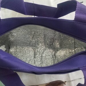 Puede incluir: Una bolsa de almuerzo aislada de color morado con cierre de cremallera y dos asas. La bolsa est&aacute; forrada con un material reflectante plateado.