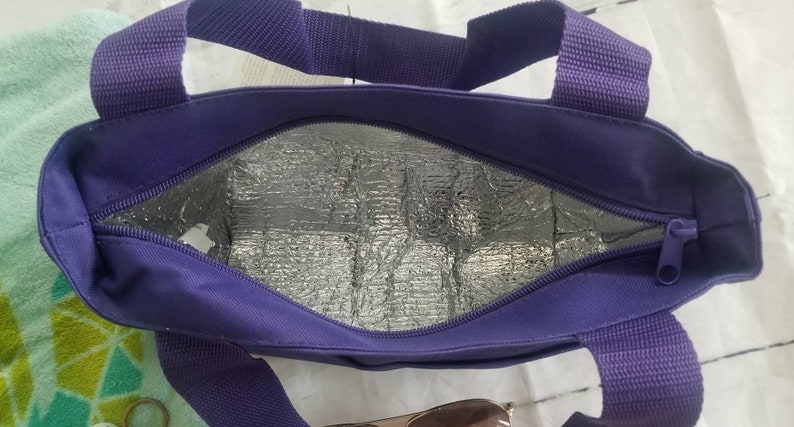 Puede incluir: Una bolsa de almuerzo aislada de color morado con cierre de cremallera y dos asas. La bolsa est&aacute; forrada con un material reflectante plateado.