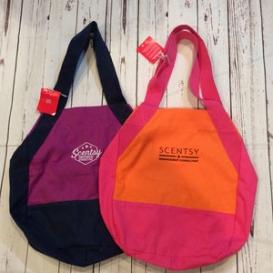 May include: Two tote bags with Scentsy branding. The bag on the left is navy blue and purple with the text "Scentsy Independent Consultant". The bag on the right is orange and pink with the text "Scentsy Independent Consultant".