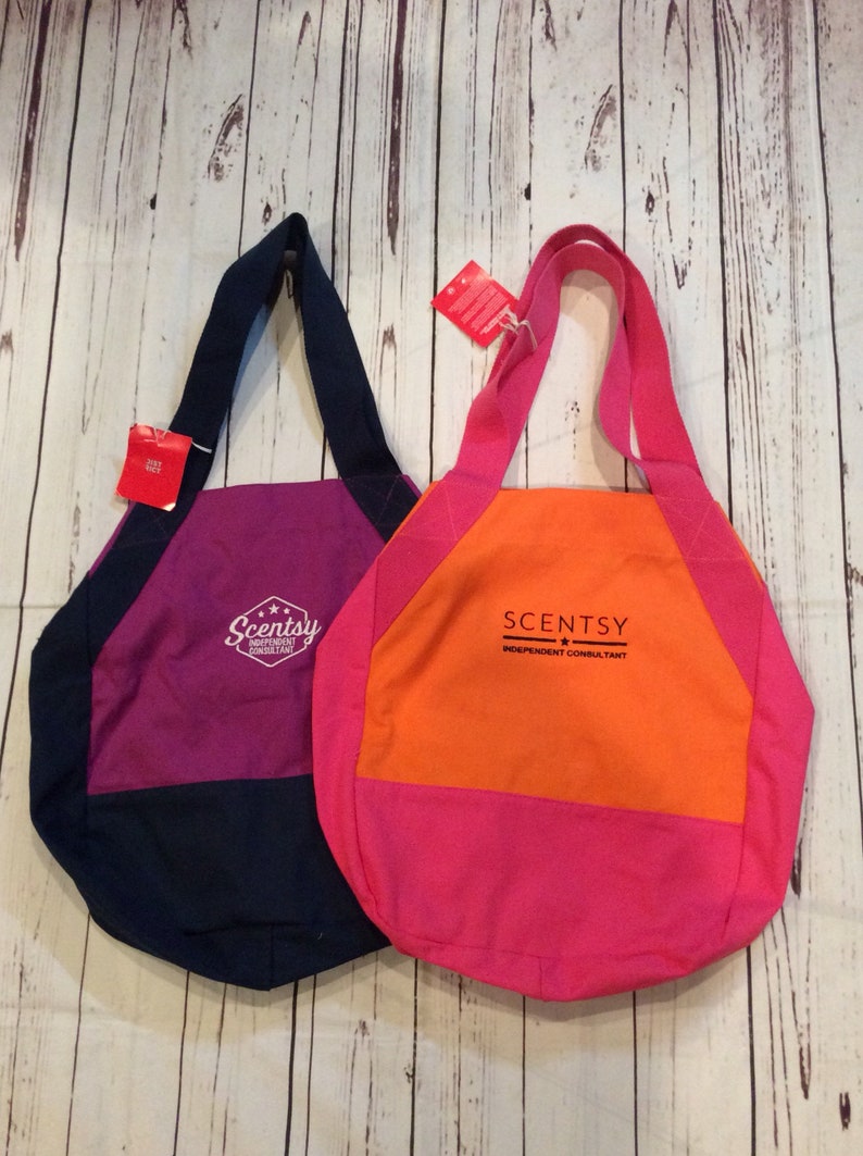 May include: Two tote bags with Scentsy branding. The bag on the left is navy blue and purple with the text "Scentsy Independent Consultant". The bag on the right is orange and pink with the text "Scentsy Independent Consultant".