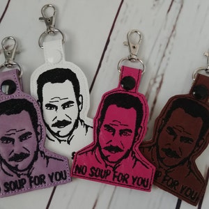 May include: Four keychains with a cartoon image of a man with a mustache and the text "No Soup for You". The keychains are in purple, white, pink, and brown.