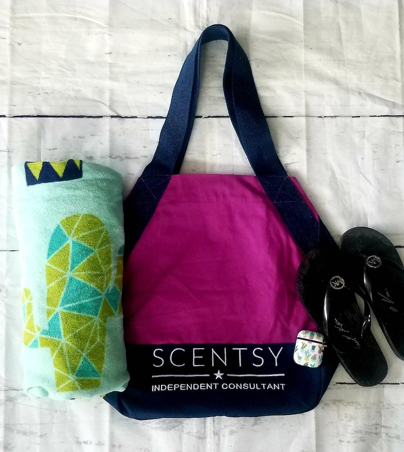 May include: A pink tote bag with navy blue straps and trim. The bag has the text "Scentsy" and "Independent Consultant" printed on the front. A green and blue geometric cactus print beach towel and black flip flops are next to the bag.