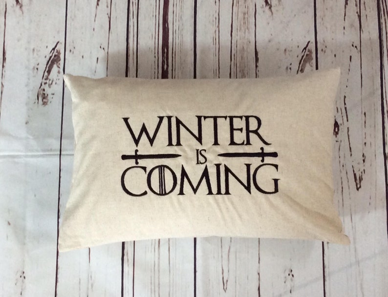 K&ouml;nnte beinhalten: Ein beiges Kissen mit dem Text "Winter is Coming" in schwarzer Stickerei. Der Text ist mit zwei Schwertern stilisiert, die sich &uuml;ber dem Wort "is" kreuzen.