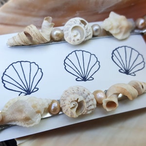 May include: Two silver hair clips decorated with seashells and pearls. The clips are on a white card with a black outline of a seashell.