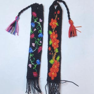 May include: Two black beaded bookmarks with colorful floral patterns and tassels. The bookmark on the left has a blue, pink, and green floral pattern. The bookmark on the right has a red and orange floral pattern.