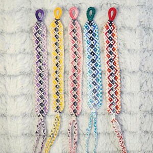May include: Five colorful friendship bracelets with a diamond pattern. The bracelets are made with different colored threads, including purple, yellow, pink, blue, and orange.