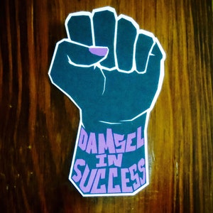 May include: A raised fist with a purple nail on the index finger. The fist is dark blue and the background is a wood grain pattern. The text "DAMSEL IN SUCCESS" is written in purple letters on the forearm.