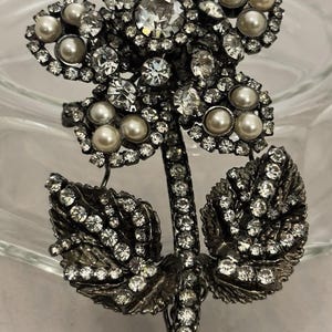 May include: A floral brooch featuring a flower design with clear crystals and faux pearls. The stem and leaves are also adorned with crystals. The brooch is set against a neutral background.