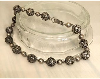 May include: A silver-toned necklace featuring alternating decorative and smooth beads. The decorative beads have a detailed swirl pattern. The necklace is displayed on a clear glass dish.