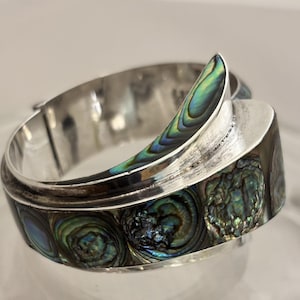 May include: A silver-toned hinged bracelet with iridescent abalone shell inlays. The bracelet features a curved design with a prominent abalone shell accent. The shell displays shades of blue, green, and purple.