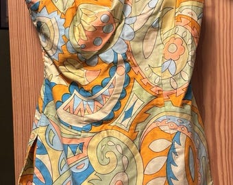 May include: A vintage-style one-shoulder swimsuit with a vibrant paisley pattern in shades of yellow, orange, blue, and green. The swimsuit is hanging on a wooden hanger against a wooden door.