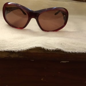 May include: A pair of red sunglasses with brown lenses. The sunglasses are on a white fabric surface.