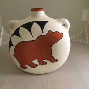 May include: A white ceramic pitcher with a brown bear silhouette and black geometric designs. The pitcher has a rounded shape and two small holes on the sides.