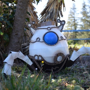 Può includere: Un robot di cartone bianco e marrone con un occhio blu e sei gambe. Il robot è in piedi in un'area erbosa con alberi sullo sfondo.