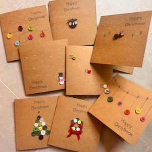 May include: A collection of eight square brown kraft paper greeting cards with the text Happy Christmas. The cards feature decorative designs made from colorful plastic buttons, including a string of lights, a pudding, a reindeer, a snowman, Ho Ho Ho text, a Christmas tree, a wreath, and hanging ornaments.