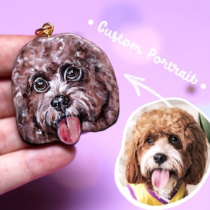 Puede incluir: Un retrato de un perro marrón y blanco pintado en un charm redondo de cerámica. El charm está colgado de una cadena dorada. El charm está siendo sostenido en la mano de una persona. El texto "Custom Portrait." está escrito en la imagen.
