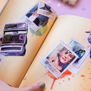 May include: Watercolor illustration of a vintage instant camera and several polaroid-style photos on a journal page. The photos feature portraits and text. A blue jay bird is perched on the page. The page has a sepia tone with watercolor paint splatters.