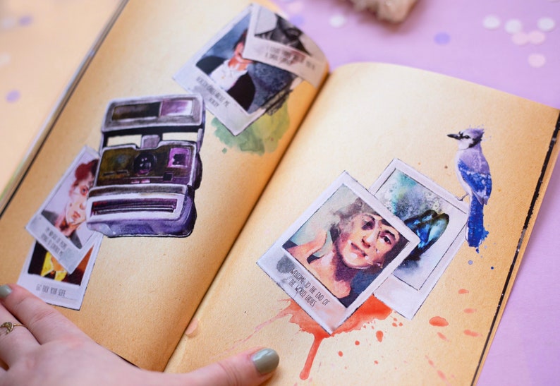 May include: Watercolor illustration of a vintage instant camera and several polaroid-style photos on a journal page. The photos feature portraits and text. A blue jay bird is perched on the page. The page has a sepia tone with watercolor paint splatters.
