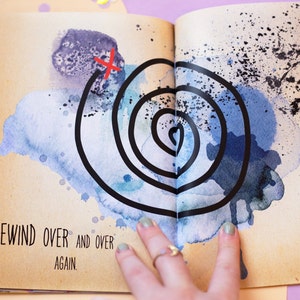 May include: Open book page with watercolor art. A black spiral design is centered on the page, with a red 'X' marking a starting point. The text 'REWIND OVER AND OVER AGAIN' is printed at the bottom of the page.
