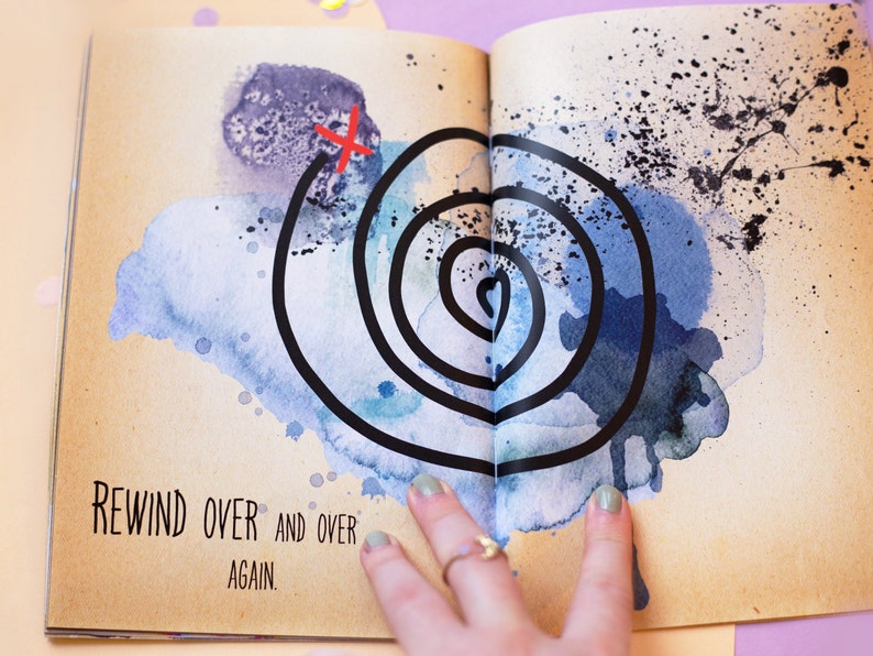 May include: Open book page with watercolor art. A black spiral design is centered on the page, with a red 'X' marking a starting point. The text 'REWIND OVER AND OVER AGAIN' is printed at the bottom of the page.