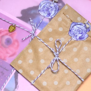 May include: A brown paper bag with white polka dots is tied with twine and a purple wax seal. The bag is sitting on a pink surface. The text on the tag reads "Happy Mail You Got This".