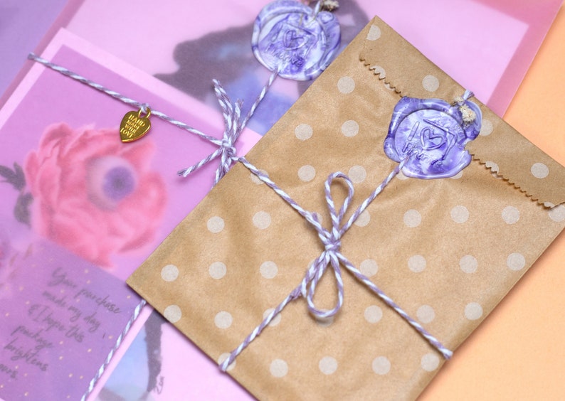 May include: A brown paper bag with white polka dots is tied with twine and a purple wax seal. The bag is sitting on a pink surface. The text on the tag reads "Happy Mail You Got This".