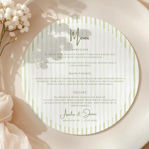 Puede incluir: Un plato blanco con una tarjeta de menú a rayas verdes. La tarjeta del menú presenta la palabra "Menú" y enumera aperitivos, platos principales y opciones de postre. Un ramo de flores blancas está a la izquierda.