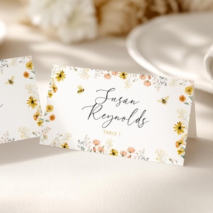 May include: Two white place cards with floral and bee accents. The cards feature the name "Susan Reynolds" in elegant script, with "Table 1" below. The cards are surrounded by yellow and peach flowers and small bees.