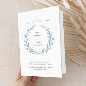 Op de afbeelding: Een witte huwelijksuitnodiging met blauwe tekst en een bloemenkransontwerp. De uitnodiging bevat de namen Eleni Stavrou en Andreas Nikolaidis, samen met de datum en locatie van de ceremonie.