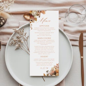 Puede incluir: Una tarjeta de menú de boda con detalles florales, que enumera los platos: Entrante, Plato principal y Postre. El menú está sobre un plato blanco con cubiertos dorados. La mesa está decorada con flores secas y un mantel rosa claro.