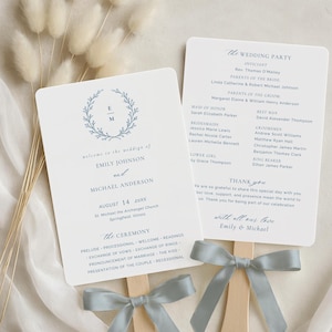 Puede incluir: Dos programas de boda en abanico con texto azul claro y una cinta a juego. El abanico izquierdo tiene una corona floral con iniciales. El abanico derecho enumera la lista de invitados. Los abanicos están sujetos a palos de madera.