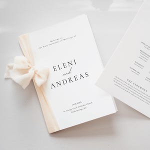 May include: Two wedding programs on a light surface. One is closed and tied with a cream ribbon. The text "ELENI and ANDREAS" is visible. The other program is open, displaying the wedding party and ceremony details.